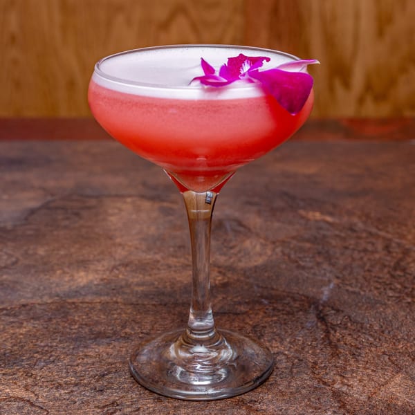 A vibrant pink cocktail with a foamy top and a purple orchid garnish, on a dark textured surface.