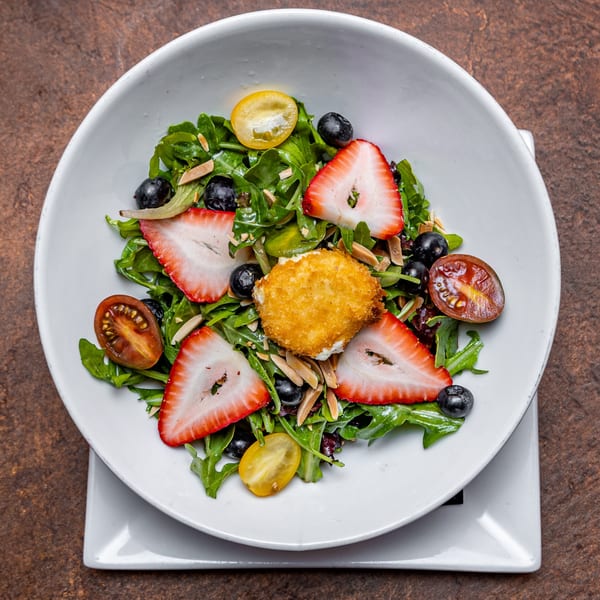 A fresh salad with strawberries, blueberries, cherry tomatoes, and fried goat cheese in a white bowl.