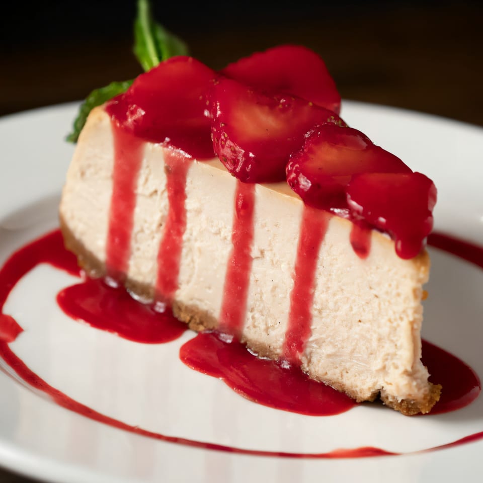 A slice of cheesecake with strawberry topping and sauce on a white plate.