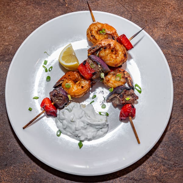 Grilled shrimp and vegetable skewers with a side of white dipping sauce and a lemon wedge.