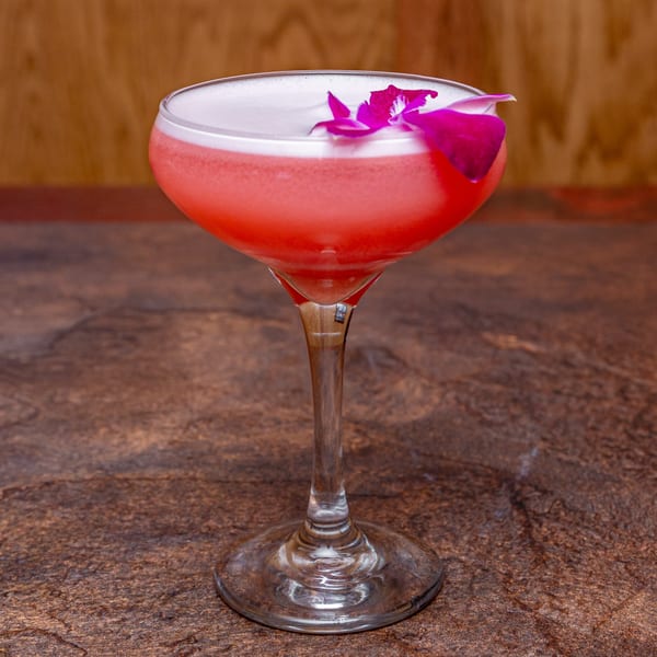 A vibrant pink cocktail with a foamy top, garnished with a purple orchid, on a dark textured surface.