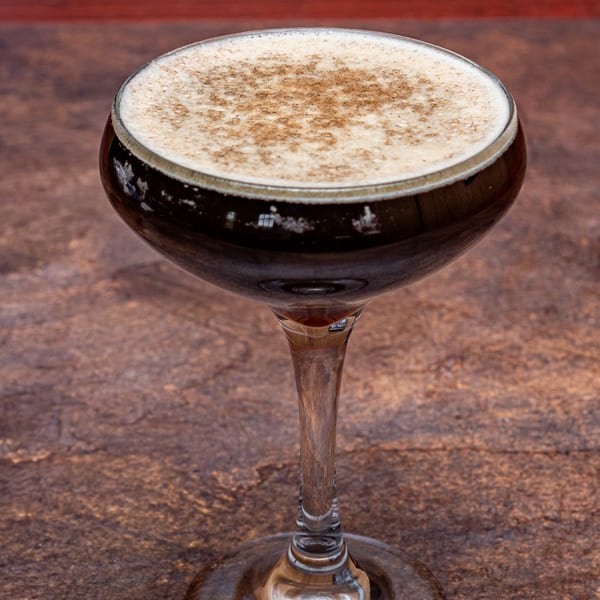 A dark cocktail with a foamy top and cinnamon dusting in a coupe glass.