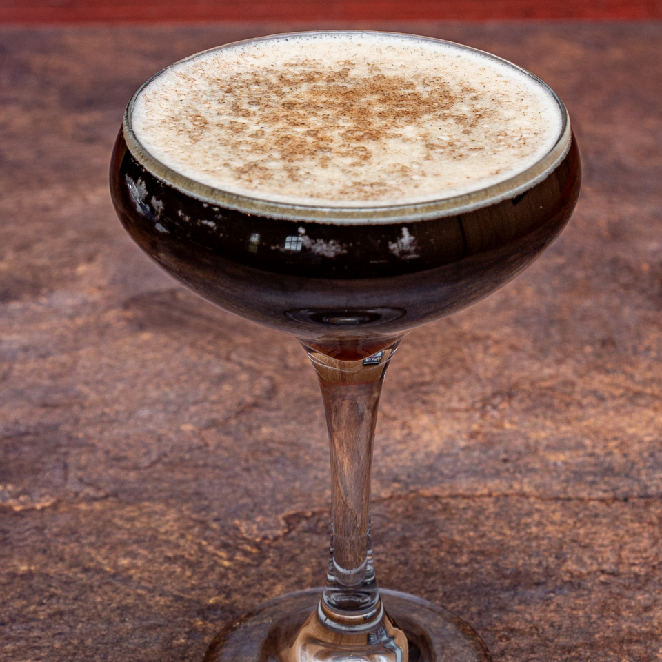 A dark cocktail with a foamy top and cinnamon dusting in a coupe glass.