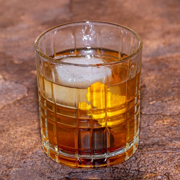 An Old Fashioned cocktail with a large ice cube, orange peel, and cherry in a textured glass.
