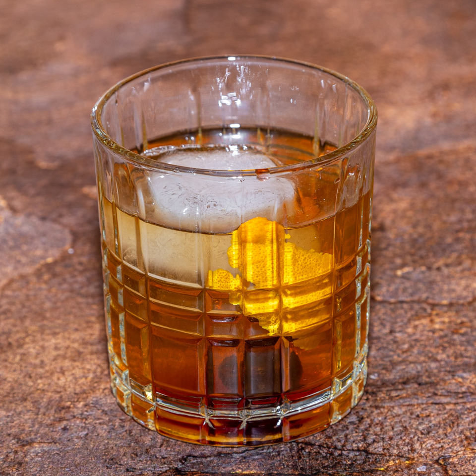 An Old Fashioned cocktail with a large ice cube, orange peel, and cherry in a textured glass.