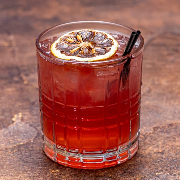 A vibrant red cocktail in a textured glass, garnished with a dried orange slice and two black straws.