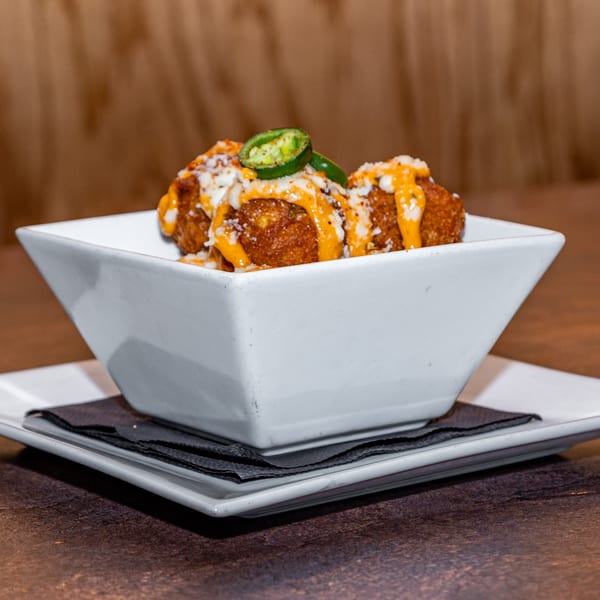 Crispy fried balls drizzled with cheese sauce and topped with sliced jalapeños in a white bowl.