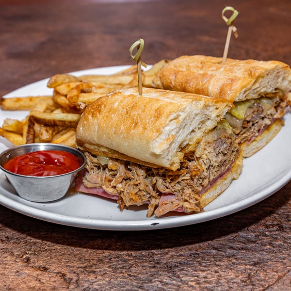 Two Cuban sandwiches with pulled pork, ham, and pickles, served with fries and ketchup on a white plate.