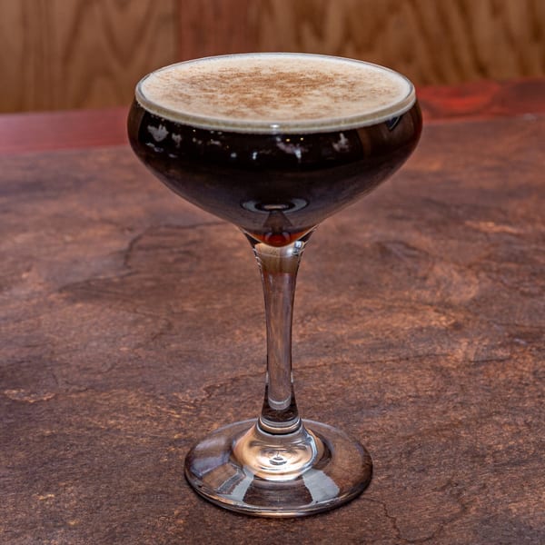 A dark cocktail with a foamy top, sprinkled with cinnamon, in a stemmed glass on a textured surface.