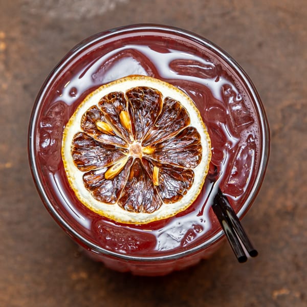 Overhead view of a red cocktail with ice, garnished with a dried orange slice and two black straws.